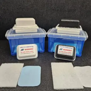 Two blue plastic containers with attached white and black handles, labeled with a warning sign. Several rectangular adhesive pads in various colors and textures are placed in front of the F-1 for 1/8″ (.32cm) to 1/4″ (64cm).