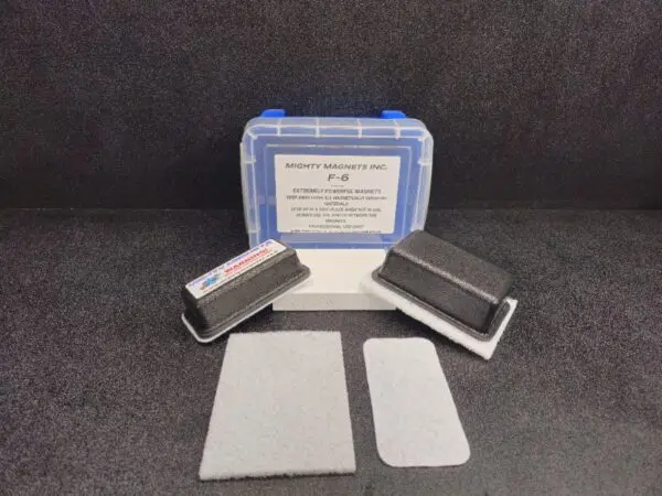 A kit from Mighty Magnets Inc. labeled F-6 for 1″ (2.5 cm) to 1-1/2″ (3.8 cm), including a clear box with a blue latch, two rectangular magnets, and two adhesive-backed metal plates. The background is a dark surface.