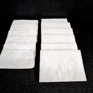 Two rows of white, rectangular, fibrous Scrub Pad (A) against a black background, one row with eight rectangular pads, the other with six rounded-edge pads.