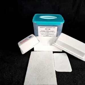 A rectangular plastic container with a blue lid labeled "Mighty Magnets Inc, FF-5 for 5/8″ (1.58 CM) TO 1-1/4″ (3.2 CM)" is surrounded by several white foam pads and magnetic cleaning tools, all placed on a black surface.