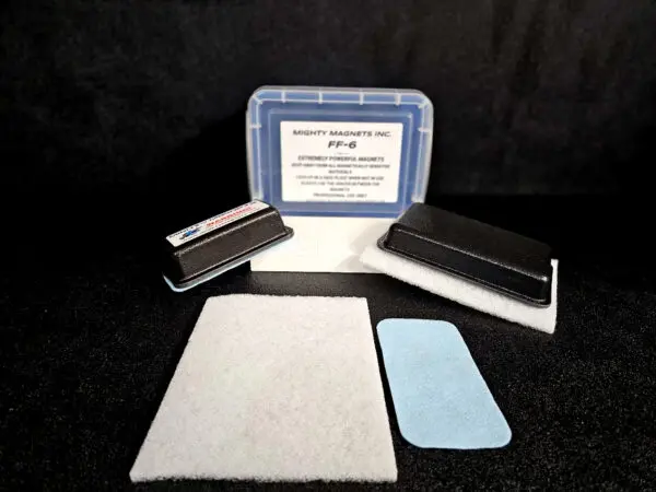 A set of cleaning supplies, including a small plastic container labeled "FF-5 for 5/8″ (1.58 CM) TO 1-1/4″ (3.2 CM)", a black scrubber pad, a white rectangular pad, and a blue rectangular pad, displayed on a black surface.